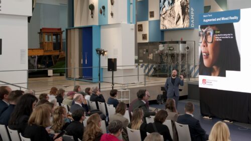 Image20251117111742 Eine Weitwinkelaufnahme eines Vortrags in einem großen, hellen Ausstellungsraum. Ein Referent steht vor einer Leinwand, auf der eine Folie zum Thema ‚Augmented and Mixed Reality‘ mit dem Gesicht einer Person, die eine Datenbrille trägt, zu sehen ist. Das Publikum sitzt in mehreren Reihen auf weißen Stühlen und blickt nach vorne. Der Raum ist mit musealen Exponaten und hohen, blau-weiß gestalteten Säulen mit Aufschriften wie ‚SEGEL‘ und ‚DAMPF‘ dekoriert.