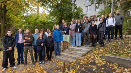 Eine große Gruppe von etwa 25 Personen posiert für ein gemeinsames Foto im Freien. Die Teilnehmenden stehen auf und neben einer flachen Betontreppe, die von herbstlichem Laub bedeckt ist. Im Hintergrund sind grüne Bäume mit ersten gelben Blättern und ein modernes, mehrstöckiges Wohngebäude zu sehen. Die Gruppe ist in herbstliche Alltagskleidung gekleidet und blickt lächelnd in die Kamera.