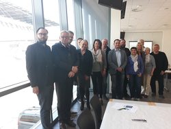 csm_20180926_Workshop_Lausitz_1_0e0313d0e3 Eine Gruppe von etwa 14 Personen posiert für ein gemeinsames Foto in einem hellen, modern gestalteten Raum. Die Gruppe steht in zwei Reihen vor einer großen Fensterfront, durch die helles Tageslicht einfällt. Alle sind in Business-Casual-Kleidung gekleidet und blicken freundlich in die Kamera. Im Vordergrund ist die Ecke eines weiß gedeckten Tisches zu sehen.