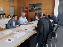 csm_20180926_Workshop_Lausitz_5_9a8096ffa4 Mehrere Personen sitzen an einem langen, weiß gedeckten Tisch in einem hellen Besprechungsraum mit Holzvertäfelung. Sie wirken konzentriert und führen Gespräche; auf dem Tisch liegen Dokumente und Flyer. Im Hintergrund sind eine Kaffeestation und ein gerahmtes Bild an der Wand zu sehen.