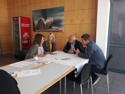 csm_20180926__Workshop_Lausitz_2_3cb781a94e Vier Personen sitzen am Ende eines langen Tisches in einem hellen Raum mit Holzvertäfelung und führen ein Gespräch. Vor ihnen auf dem Tisch liegen Notizen und eine Kaffeetasse. Im Hintergrund ist ein roter Getränkekühlschrank sowie ein großes, gerahmtes Bild an der Wand erkennbar.
