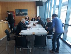 csm_20180926__Workshop_Lausitz_3_db6788c189 Eine Szene aus einem Workshop oder einer Besprechung: Während ein Teil der Gruppe am Tisch sitzt, steht eine Person rechts im Bild und beugt sich leicht vor.